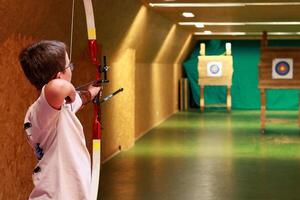 Boy with a bow and an arrow aiming at a target at an inside archery shooting range. photo