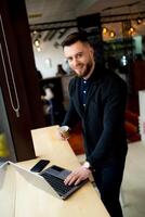 Man with nice smile stands near laptop in coffee shop. Fashion clothes on a handsome man. Coffee break in the middle of the day. Closeup. photo