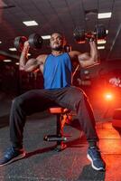 Cute african american doing workout with two dumbbells while sitting on bench in gym. View from below. photo