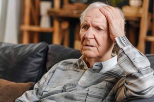 Elderly Man Sitting on Couch with Hand on Head in Thoughtful Pose at Home, Wearing Gray Sweater, Indoor, Unposed Moment, Senior Lifestyle and Emotions, Realistic Representation of Everyday Life photo