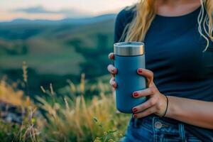 Woman Holding Travel Mug in Mountainous Landscape During Golden Hour, Scenic Outdoor Adventure with Blue Stainless Steel Insulated Container, Grass and Hills in Background photo