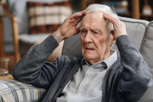 Elderly Man Looking Worried at Home, Holding Head in Hands, Wearing Sweater, Sitting on Couch, Interior Background photo