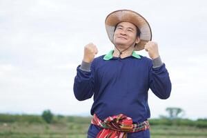 Handsome Asian man farmer stands at agricultural land, wears hat, Thai loincloth on waist, raises fists up with excited feeling or success. Concept, agriculture occupation. photo