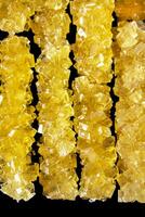 The sticks are made of sugar and are arranged in a line. A row of yellow crystals are lined up on a black surface. The crystals are all different sizes and shapes photo