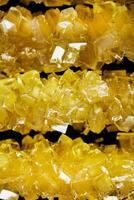 The sticks are made of sugar and are arranged in a line. A row of yellow crystals are lined up on a black surface. The crystals are all different sizes and shapes photo