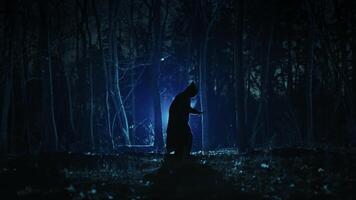 A dark figure in a cloak with a hood in a sinister night forest. Fantasy demon or witcher in the night. video
