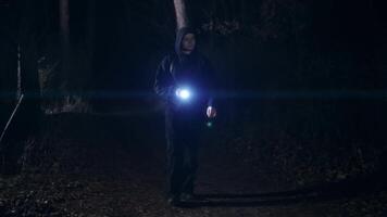A young guy with a backpack on his back walks through a forest or park at night. A man in a hoodie and with a flashlight in his hand video