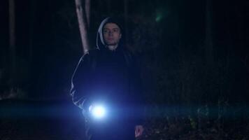 A young guy with a backpack on his back walks through a forest or park at night. A man in a hoodie and with a flashlight in his hand video