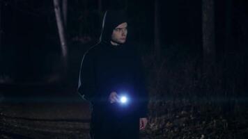 A frightened young guy walks through the night forest with a flashlight and looks around. The dark night time is frightening with the unknown. video