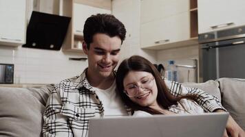 Young couple sitting together on the couch at home, watching something on a laptop, hugging each other and smiling. Concept of happiness and harmony in a relationship video