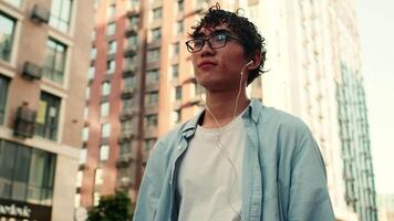 Young asian curly man in casual clothes and glasses walking in wired headphones with a smartphone and smiling on a city street on a sunny day with multi-storey buildings as a backdrop video