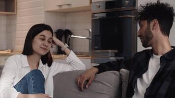 Young man in casual clothes telling something to his smiling girlfriend while sitting on the couch in a cozy kitchen at home. Concept of happiness and harmony in a couple relationship video