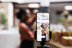 The eco-conscious owner promotes zero waste with reusable packaging. Social media vlogger films using a smartphone and tripod. Vibrant shelves and a camera capture the moment. Focus on the foreground. photo