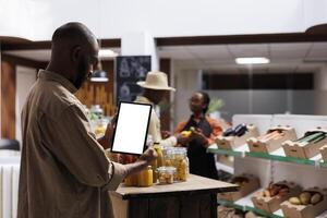 African American male customer vertically grasping a digital tablet with an isolated chromakey mockup template. The store features reusable packaging and sustainable products on wooden shelves. photo