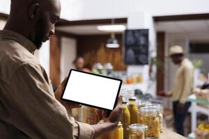 An African American guy consumer holds a phone tablet horizontally with an isolated chromakey mockup template. On wooden shelves, the store sells fresh, eco friendly products. photo