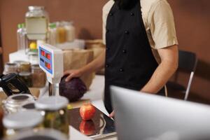 Caucasian store worker in black apron, scans and checks a red apple on weight scale, while he holds a big purple cabbage in his hand. Salesman measuring natural organic produce at cashier desk. photo