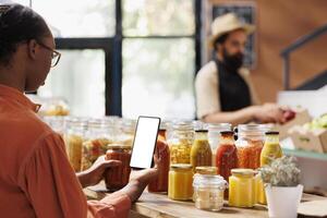 Black woman grasping mobile device with white screen while browsing the wooden counter for bio food products. Female client searching for organic items while holding smartphone with mockup template. photo