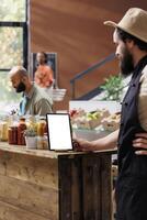 Male storekeeper wearing a hat and black apron glances at tablet with white screen placed vertically on table. Caucasian vendor using device with blank copyspace mockup template. photo