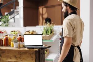 Local shop vendor looks at laptop showing isolated chromakey template near shelves filled with fresh food supplies. Caucasian seller uses minicomputer with white screen display. photo