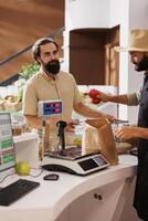 Young storekeeper attending to a customer at the cashier desk. Products are packed into a brown paper bag for the caucasian man behind the counter by vendor selling things with an apron and a hat. photo