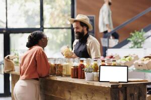 Horizontally positioned tablet with a blank chromakey mockup template near glass jars. While the shopkeeper speaks with the customer, a smart device on the counter displays an isolated white screen. photo