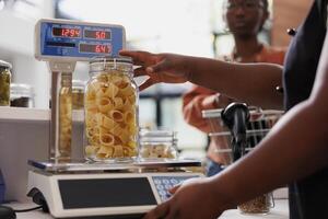 Closeup of african american vendor using digital scale to measure weight of fresh organic product in glass container. Detailed view of merchant weighing jar of bio pasta while client waits at counter. photo