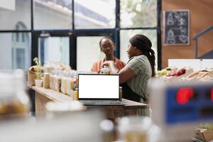 Digital device with blank copy space mockup template is set up on wooden counter as vendor assists customer. An isolated white screen displayed on laptop for eco friendly bio store commercial. photo