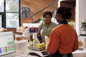 Smiling black lady receiving assistance from African American vendor at the checkout counter with her locally grown produce. Customer at cashier desk in zero waster store purchasing organic products. photo