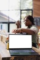 At supermarket, African American client is guided by a vendor while prioritizing sustainable and organic items. Simultaneously, laptop screen shows blank chromakey for advertising bio food content. photo