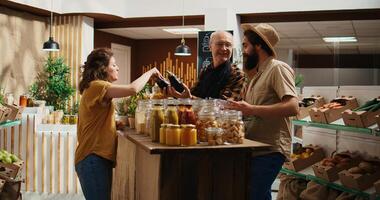 Senior vendor assisting clients in eco friendly shop with ethical sourced chemicals free food items. Couple in local store chatting with trader about pantry staples bulk products, zoom out photo