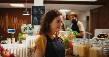 Portrait of smiling woman working in zero waste organic food supermarket, adding pantry staples on shelves. Cheerful employee restocks local grocery store with bulk products in reusable jars photo