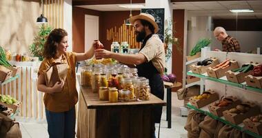 Client talking with seller in organic food shop, inquiring about products ethical sourcing and asking for recommendations. Woman in low carbon footprint store chatting with trader about pantry staples photo