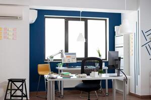 Empty business office room in modern open space workplace. Desk with computer and supplies in commercial workplace with big window. Workspace furniture inside startup with blue and white wall. photo