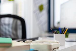 Closeup of phone on desk in empty business office. Startup workspace furniture in indoors work location. Modern commercial workplace design with telephone on white table with supplies. photo