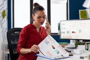Business woman explaining charts on the phone. Entrepreneur talking on the telephone sitting at startup desk with desktop computer. Successful employee in red shirt explaining data to manager. photo