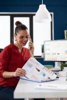 Smiling entrepreneur talking on the telephone explaining charts. Successful manager happy with business results in startup office. Employee in red shirt working at desk with clipboard and computer. photo
