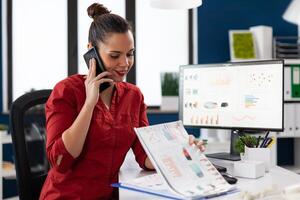 Entrepreneur explaining charts on the mobile phone. Smiling employee talking on the smartphone sitting at startup desk with desktop computer. Successful manager in red shirt happy with business results. photo