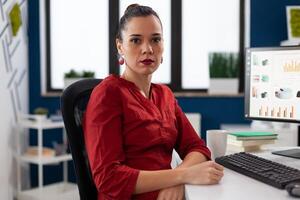Portrait of businesswoman sitting at desk in startup office. Professional entrepreneur looking confident in business workspace. Successful employee in red shirt posing in front of desktop computer. photo