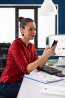 Manager using smartphone in startup business office. Businesswoman in red shirt texting sitting at desk. Company employee browsing internet on the phone in front of desktop computer. photo