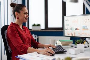 Entrepreneur looking at charts on computer screen in startup office. Smiling businesswoman working on reports at desk. Successful employee in red shirt using desktop pc to analyze business data. photo