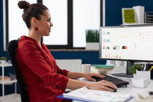 Successful businesswoman using desktop computer to analyze business data. Smiling entrepreneur looking at charts on pc screen in startup office. Employee in red shirt working on reports at desk. photo