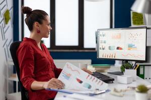 Businesswoman with clipboard sitting at desk in startup business office. Smiling employee in red shirt comparing charts. Successful entrepreneur looking at desktop computer to analyze business data. photo