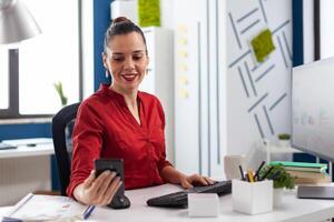 Businesswoman in videocall conference using smartphone. Manager using mobile phone in startup business office. Employee in red shirt sitting at desk browsing internet in front of desktop computer. photo