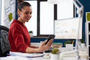 Startup employee explaining business results in videocall. Manager in red shirt sitting at desk in office discussing charts in online call. Businesswoman working in front of desktop computer. photo