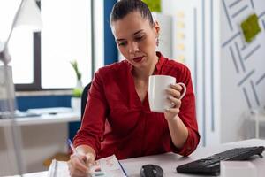 Startup manager holding cup writing on clipboard with chart. Entrepreneur in red shirt with pen in one hand working at desk in startup office. Focused employee analyzing small business data. photo