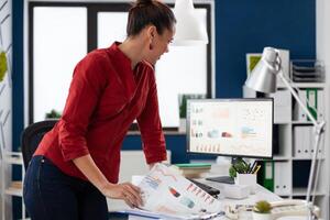 Entrepreneur leaning over desk looking at charts on desktop computer screen. Businesswoman standing comparing business data with clipboard. Employee in red shirt looking at pc monitor. photo