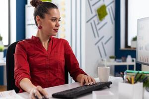 Content businesswoman reading chart on computer screen. Startup owner with clipboard happy with business results. Entrepreneur in red shirt typing on pc keyboard while looking at display. photo