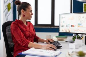 Businesswoman sitting at desk in startup office typing business data. Smiling startup manager in red shirt looking at charts on computer monitor. Entrepreneur with clipboard analyzing graph and data. photo