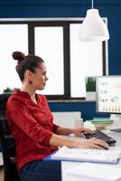 Startup owner with clipboard working at office desk with overhead lamp. Entrepreneur analyzing business chart on computer screen. Businesswoman in red shirt typing data on keyboard. photo