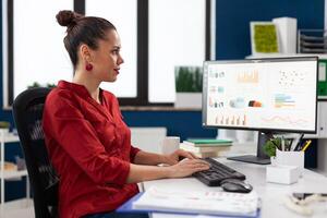 Startup manager in red shirt looking at charts on computer monitor. Smiling small business owner typing on keyboard at desk in start up office. Employee with clipboard analyzing graph and data. photo